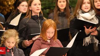 Ein Chor junger Mädchen, einige mit Brille und Schals, halten Notenblätter und singen in einer Freiluftumgebung.