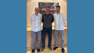 Drei Männer stehen in einer Küche zusammen, zwei in Kochuniformen und einer in Freizeitkleidung, lächeln und posieren für ein Foto vor einem dekorativen Gemälde.