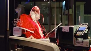 Ein Mann in einer Santa-Claus-Verkleidung sitzt in einem Fahrzeug, trägt eine rote Kostüm mit weißem Bart und Brille. Er ist von Bürogeräten umgeben, und ein Fernseher ist an der Wand montiert.