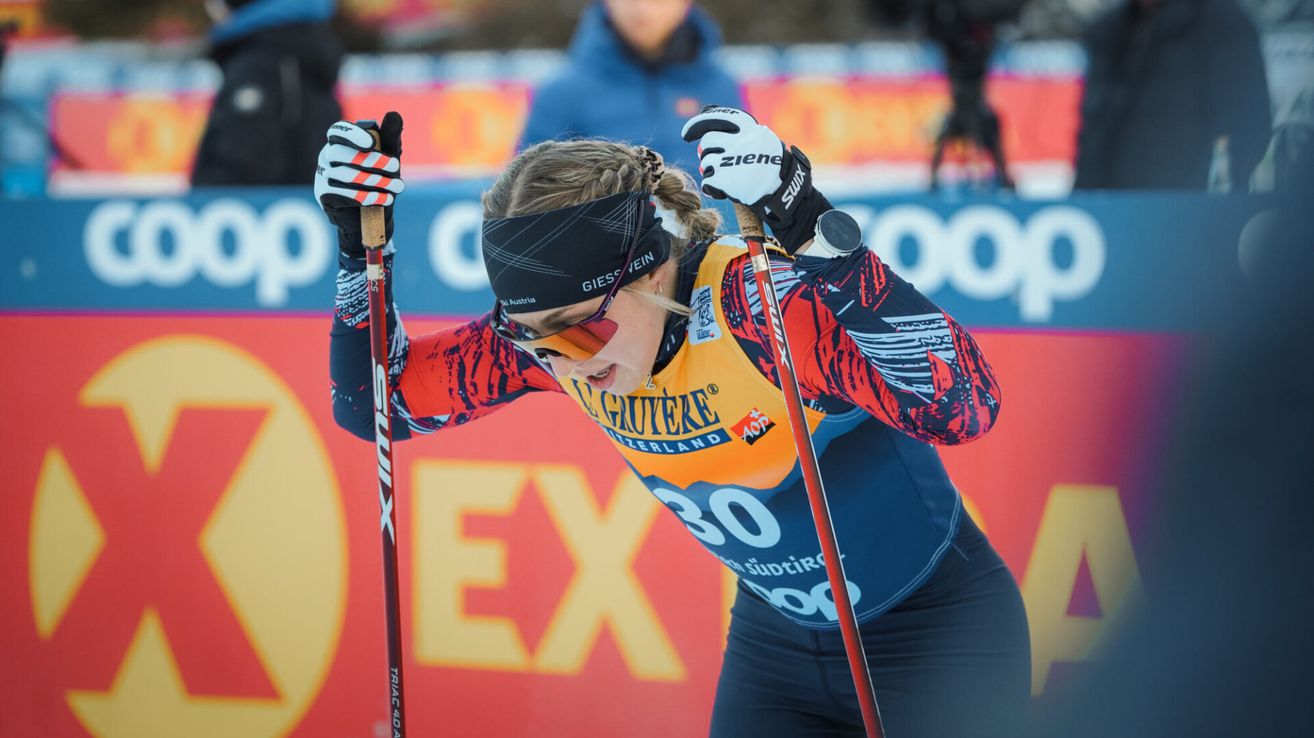 Eine Langläuferin in einem gelben und blauen Anzug mit den Aufschriften 'L'GUVIZEKE' und 'SCHWEIZLAND' steht auf dem Schnee, hält Skistöcke und schaut nach vorne. Hinter ihr ist ein Banner mit gelben und blauen Farben zu sehen.