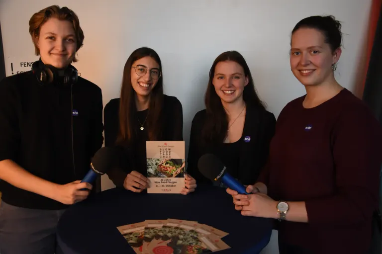 Vier Personen, zwei Männer und zwei Frauen, stehen um einen Tisch herum und halten Mikrofone. Sie lächeln und scheinen sich auf ein Interview vorzubereiten. Ein Büchlein mit dem Titel 'Slow Food Tage' liegt auf dem Tisch.