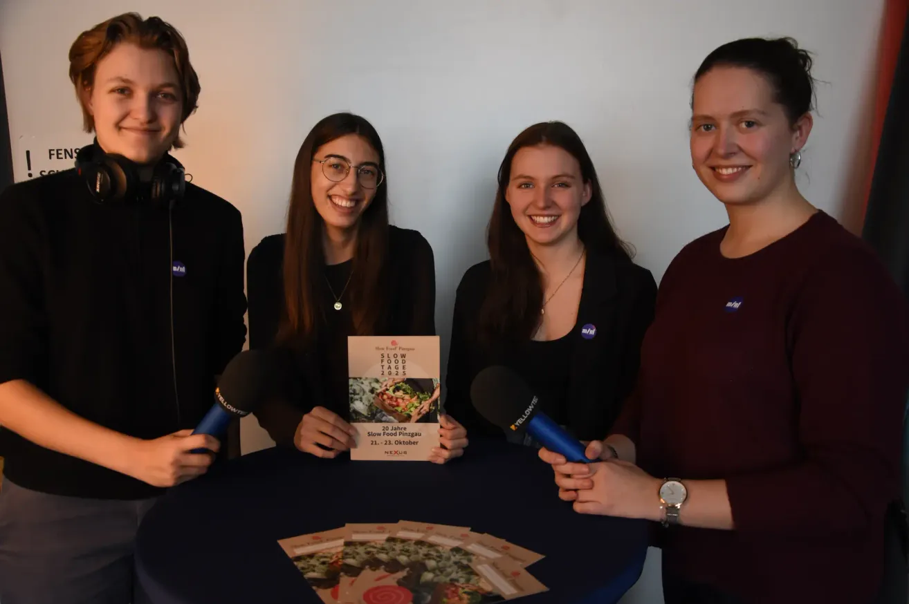Vier Personen, zwei Männer und zwei Frauen, stehen um einen Tisch herum und halten Mikrofone. Sie lächeln und scheinen sich auf ein Interview vorzubereiten. Ein Büchlein mit dem Titel 'Slow Food Tage' liegt auf dem Tisch.