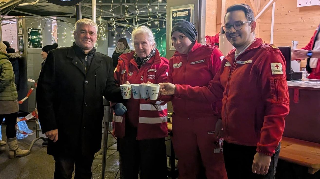 Vier Personen in roten Uniformen stehen lächelnd nebeneinander und halten Tassen in der Hand. Eine Frau trägt einen weißen Schal. Hinter ihnen steht eine Person, und Lichter hängen von der Decke.