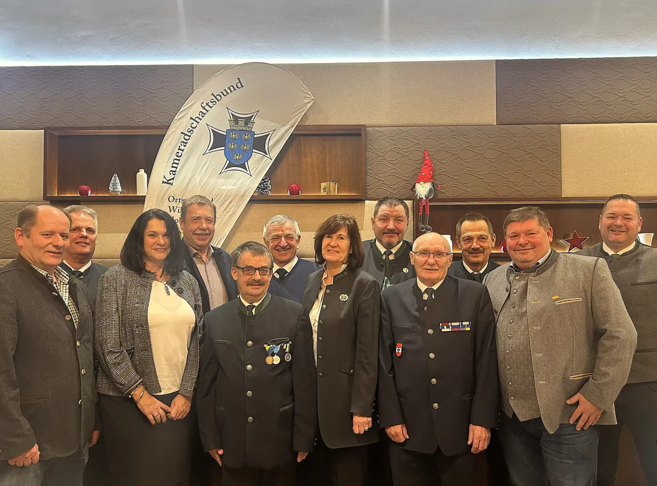 Eine Gruppe von Menschen, möglicherweise ein Team, posiert für ein Foto in einem Konferenzraum. Dahinter sind ein Banner und ein Regal mit verschiedenen Gegenständen zu sehen. Die Gruppe besteht aus Männern und Frauen, alle formal gekleidet.