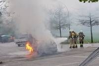 Zwei Feuerwehrleute löschen einen Autobrand auf einem Parkplatz, wobei Rauch aus dem Fahrzeug aufsteigt. Ein LKW ist in der Nähe geparkt.