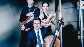 Eine Gruppe von vier Musikern, zwei Männern und zwei Frauen, in formeller Kleidung, posiert für ein Foto, während sie Saiteninstrumente halten, darunter Violinen und ein Cello.