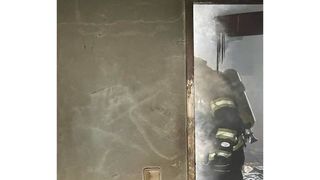 Ein Feuerwehrmann betritt ein Gebäude durch eine offene Tür, hält einen Feuerlöscher in der Hand und trägt Schutzausrüstung. Rauch ist aus dem Raum sichtbar.