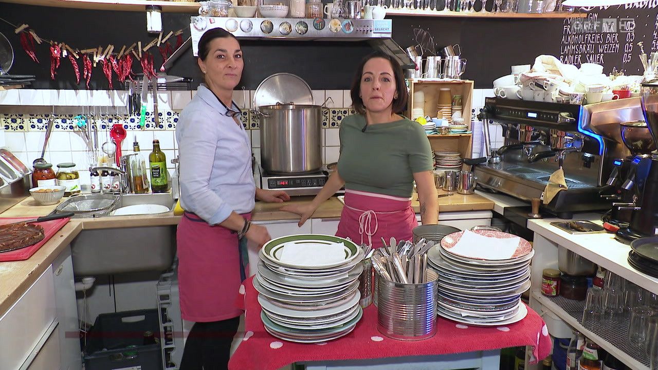 Zwei Frauen in Schürzen in einer Küche, eine mit einem Kochtopf auf dem Herd, die andere mit einem Stapel Teller auf einem Tisch.
