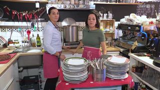 Zwei Frauen in Schürzen in einer Küche, eine mit einem Kochtopf auf dem Herd, die andere mit einem Stapel Teller auf einem Tisch.