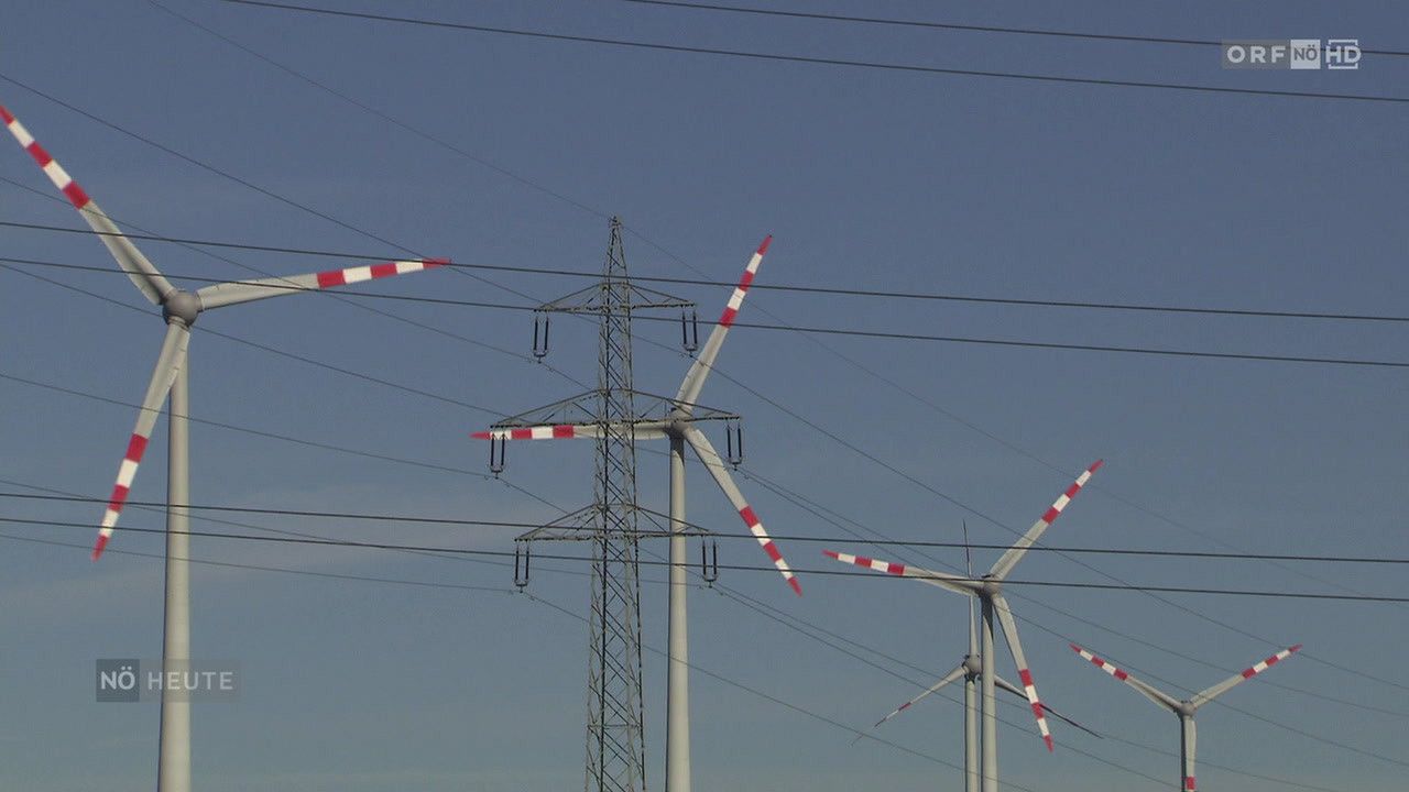 Drei Windturbinen mit roten und weißen Rotorblättern sind durch mehrere Drähte verbunden, vor einem blauen Himmel. Ein hoher Metallmast steht zwischen zwei von ihnen.