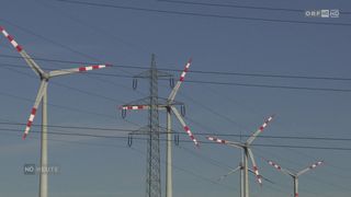 Drei Windturbinen mit roten und weißen Rotorblättern sind durch mehrere Drähte verbunden, vor einem blauen Himmel. Ein hoher Metallmast steht zwischen zwei von ihnen.