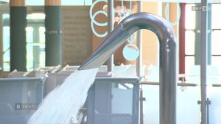 Ein Edelstahlhahn spritzt Wasser in einen Pool, mit einem dekorativen Design an der Wand dahinter.