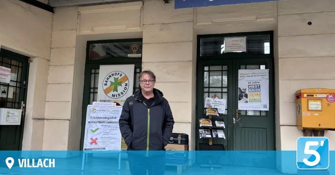 Ein Mann steht vor einem Gebäude mit einem Schild, das 'Bahnhofs Mission' und ein anderes Schild mit 'Kleiderbgabe' steht. Er trägt eine Brille und eine Jacke. Es gibt Plakate und einen gelben Briefkasten am Gebäude. Der Wasserzeichen liest 'VILLACH'.