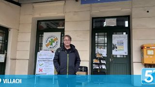 Ein Mann steht vor einem Gebäude mit einem Schild, das 'Bahnhofs Mission' und ein anderes Schild mit 'Kleiderbgabe' steht. Er trägt eine Brille und eine Jacke. Es gibt Plakate und einen gelben Briefkasten am Gebäude. Der Wasserzeichen liest 'VILLACH'.