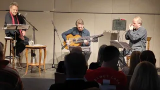 Ein Mann spielt Gitarre, während ein anderer eine Mundharmonika vor einem Publikum spielt. Sie stehen auf einer Bühne mit einem Holztisch und Mikrofonen. Das Publikum sitzt vor ihnen.