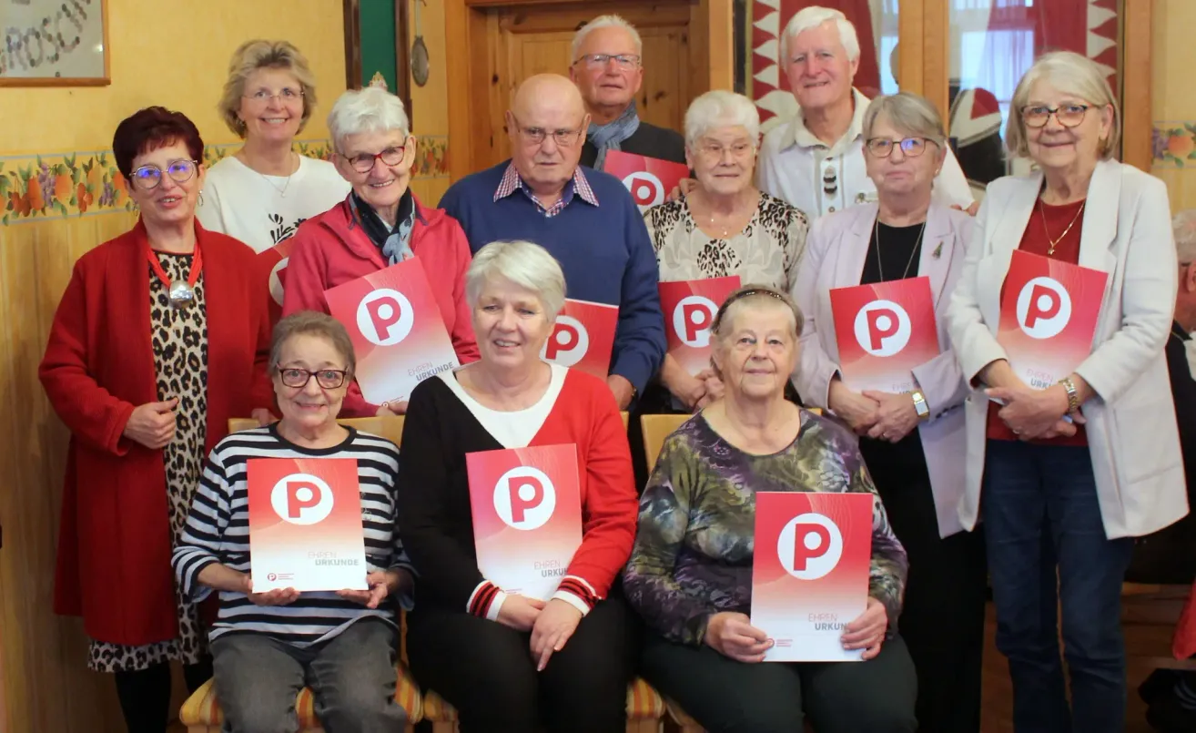 Eine Gruppe älterer Personen mit Brillen posiert für ein Foto. Sie halten ein rotes Plakat mit einem weißen Buchstaben P. Der Mann im blauen Hemd schaut etwas an. Hinter ihnen ist eine Holzwand mit einem hängenden Ornament. Der Raum ist schwach beleuchtet.