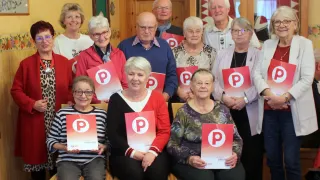 Eine Gruppe älterer Personen mit Brillen posiert für ein Foto. Sie halten ein rotes Plakat mit einem weißen Buchstaben P. Der Mann im blauen Hemd schaut etwas an. Hinter ihnen ist eine Holzwand mit einem hängenden Ornament. Der Raum ist schwach beleuchtet.