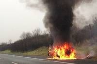 Ein Fahrzeug brennt am Straßenrand, wobei schwarzer Rauch aufsteigt. Die Straße ist leer, und es gibt Büsche und Bäume am Straßenrand.