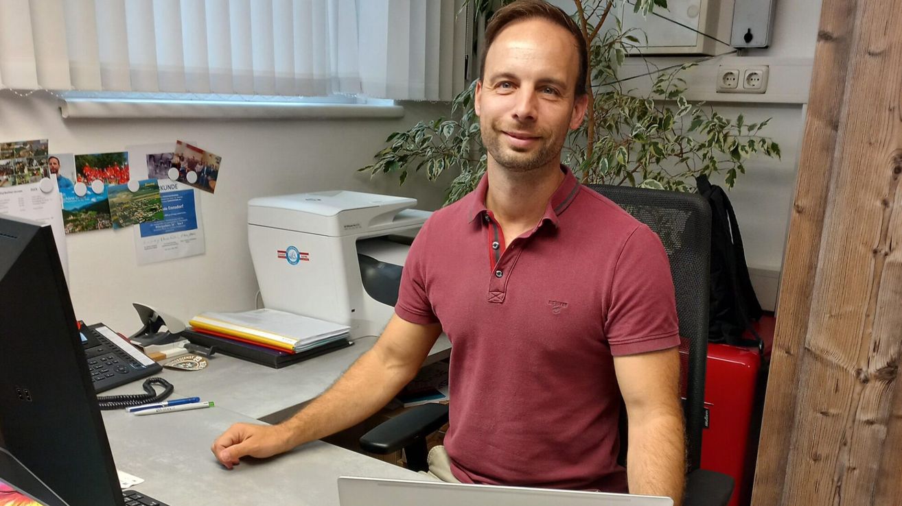 Ein Mann in einem weinroten Polohemd sitzt mit einem Laptop an einem Schreibtisch neben einem Drucker und einer Pflanze. Er lächelt in die Kamera.