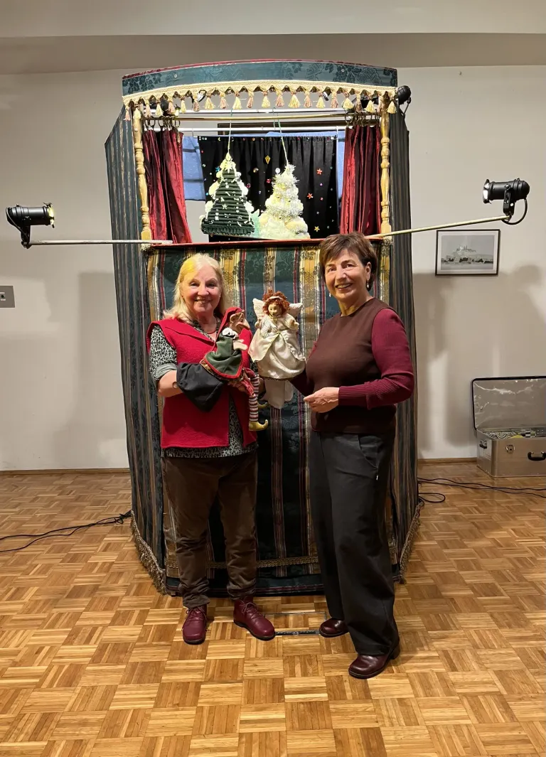 Zwei Frauen stehen vor einer Bühnenkulisse, eine hält Puppen und lächelt. Hinter ihnen befindet sich ein Weihnachtsbaum, ein Rahmen und eine Tasche.