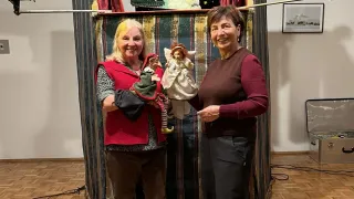 Zwei Frauen stehen vor einer Bühnenkulisse, eine hält Puppen und lächelt. Hinter ihnen befindet sich ein Weihnachtsbaum, ein Rahmen und eine Tasche.