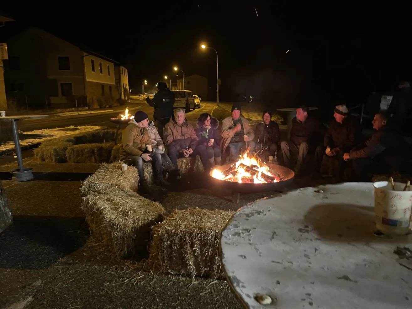 Eine Gruppe von Menschen sitzt nachts um ein Lagerfeuer in einem ländlichen Gebiet. Sie sind warm gekleidet und haben Tassen in den Händen. Einige tragen Mützen und Schuhe. Im Hintergrund sind Straßenlaternen und ein Van zu sehen.