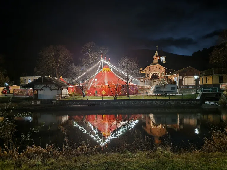 Ein Zirkuszelt ist nachts in der Nähe eines Sees beleuchtet. Das Zelt hat rote und gelbe Streifen, und ein gelber Stern ist auf der Spitze. Der See reflektiert die Lichter des Zeltes.