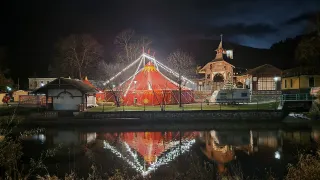Ein Zirkuszelt ist nachts in der Nähe eines Sees beleuchtet. Das Zelt hat rote und gelbe Streifen, und ein gelber Stern ist auf der Spitze. Der See reflektiert die Lichter des Zeltes.