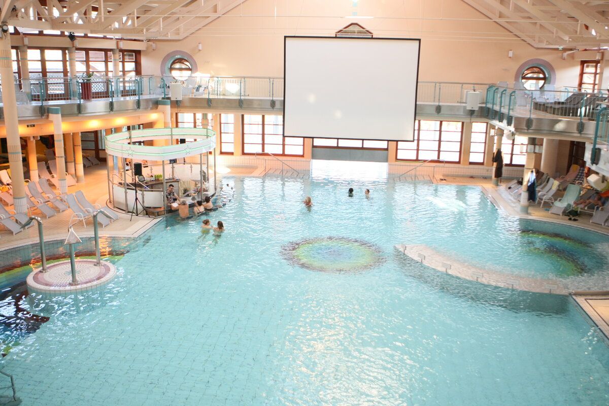 Überdachtes Schwimmbad mit Schwimmenden. Große Projektionsleinwand über dem Pool. Erhöhte Plattformen an den Seiten.