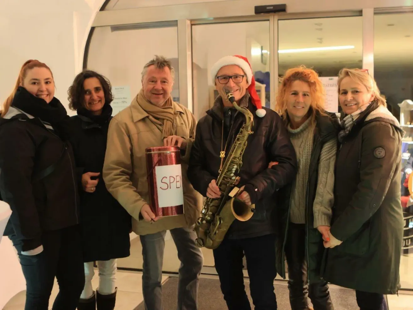 Fünf Menschen posieren für ein Foto. Zwei Männer tragen Weihnachtsmützen, einer hält eine Saxophon und der andere eine Spendendose. Die Frauen tragen Winterkleidung. Hinter ihnen ist eine Glastür.