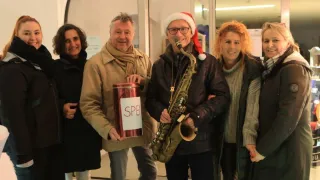 Fünf Menschen posieren für ein Foto. Zwei Männer tragen Weihnachtsmützen, einer hält eine Saxophon und der andere eine Spendendose. Die Frauen tragen Winterkleidung. Hinter ihnen ist eine Glastür.