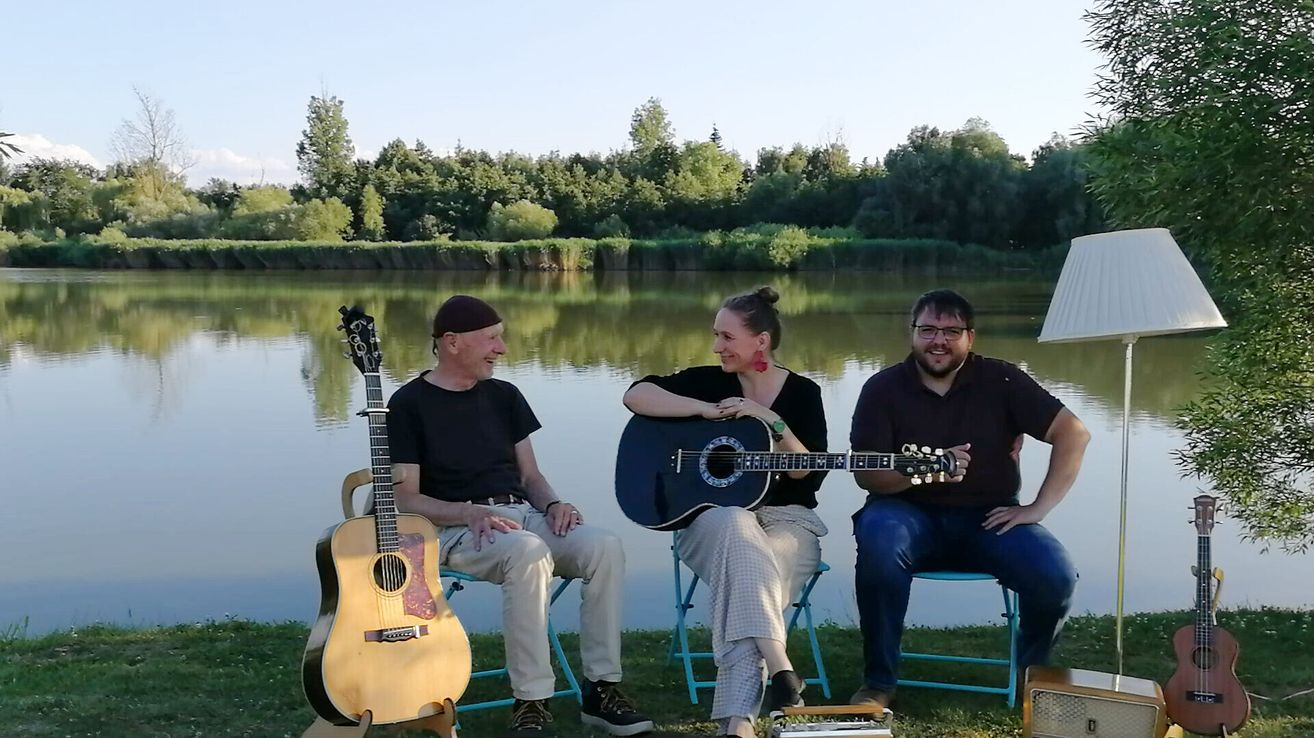 Drei Personen sitzen auf Stühlen am See und halten Gitarren. Der Mann links trägt eine Mütze und Turnschuhe, während die anderen beiden lächeln. Die Frau in der Mitte trägt Ohrringe. Der See ist von Bäumen umgeben.