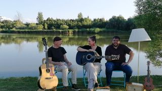 Drei Personen sitzen auf Stühlen am See und halten Gitarren. Der Mann links trägt eine Mütze und Turnschuhe, während die anderen beiden lächeln. Die Frau in der Mitte trägt Ohrringe. Der See ist von Bäumen umgeben.
