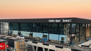 Eine Luftaufnahme des Terminals 3 des Flughafens Frankfurt während des Sonnenuntergangs. Das Terminal hat ein modernes Design mit großen Glasfenstern. Im Vordergrund sind Bauarbeiten zu sehen.