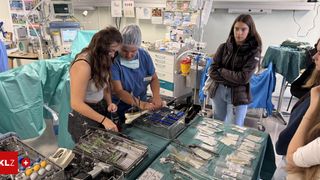 Drei Frauen sind in einem Operationssaal. Eine Frau trägt eine blaue Uniform und eine Kappe, eine andere ein graues Oberteil und schwarze Hose, und die dritte eine braune Jacke und Jeans. Sie bereiten medizinische Instrumente auf einem Tisch vor.