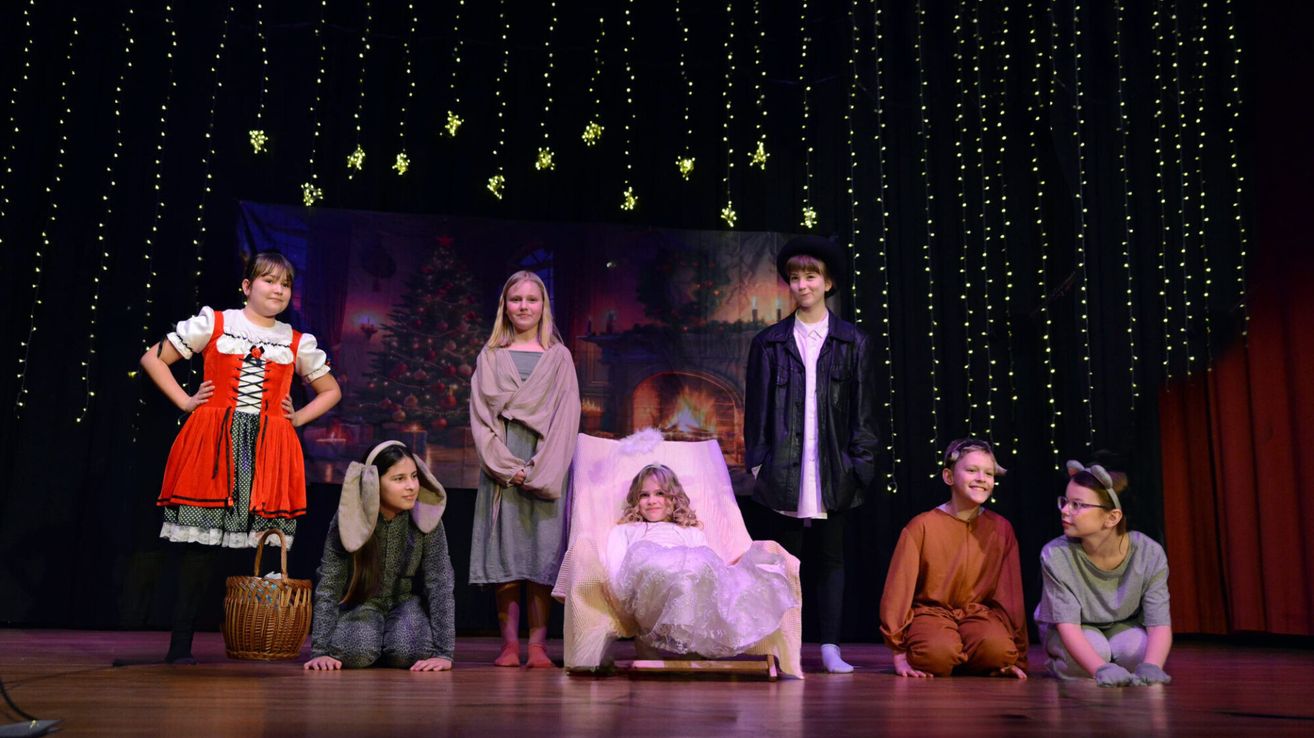 Eine Gruppe von Kindern steht auf der Bühne, in Kostümen gekleidet, mit einem Hintergrund von einem Weihnachtsbaum und Kamin. Sie spielen wahrscheinlich ein Theaterstück.