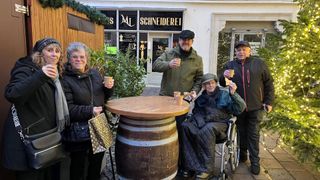 Vier Personen stehen um einen Holzfass herum, lächeln und halten Tassen. Hinter ihnen ist ein Geschäft mit dem Schild 'ML SCHNEIDEREI' zu sehen. Das Geschäft hat Glasfenster und eine Pflanze davor.
