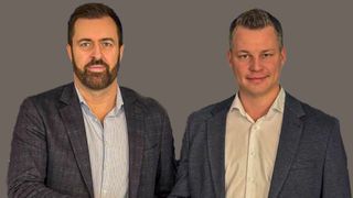 Zwei Männer in Anzügen und hellen Hemden posieren für ein Bild vor einem schlichten grauen Hintergrund. Der Mann auf der linken Seite hat einen Bart, während der Mann auf der rechten Seite einen glatt rasierten Gesichtsausdruck hat.