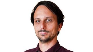 Ein Mann mit kurzem Bart und Schnurrbart trägt ein weinrotes Hemd und lächelt.