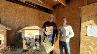 Zwei Männer stehen neben einem Miniaturhaus, einer hält eine Figur. Das Haus hat eine Brücke und Figuren davor. Der Hintergrund ist hölzern.