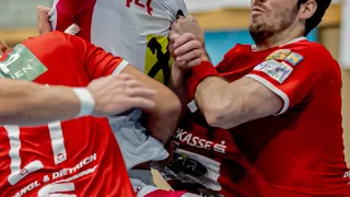 Drei Spieler sind in einem intensiven Handballspiel. Zwei Männer ringen, während die Frau hinter ihnen zusieht. Der Mann auf der rechten Seite hat die Nummer 244 auf seinem Trikot.