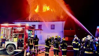 Feuerwehrleute löschen ein Feuer auf einem Haus bei Nacht. Ein Feuerwehrwagen sprüht Wasser auf das brennende Dach.
