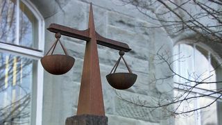 Eine hölzerne Waage-Skulptur steht vor einem Gebäude aus Stein mit weißen Fenstern und kahlen Ästen. Die Waage balanciert mit zwei Körben auf beiden Seiten.