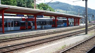 Ein Bahnhof mit einem Mann, der in der Nähe eines Zuges steht. Der Bahnhof hat ein blaues Schild mit der Aufschrift 'Bruck an der Mur'. Es gibt zwei Gleise und Bänke auf dem Bahnsteig.