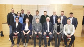 Eine Gruppe von Personen posiert für ein Foto in einem Raum. Sie sitzen auf Stühlen und lächeln alle. Der Raum hat Holzpaneele an der Wand.