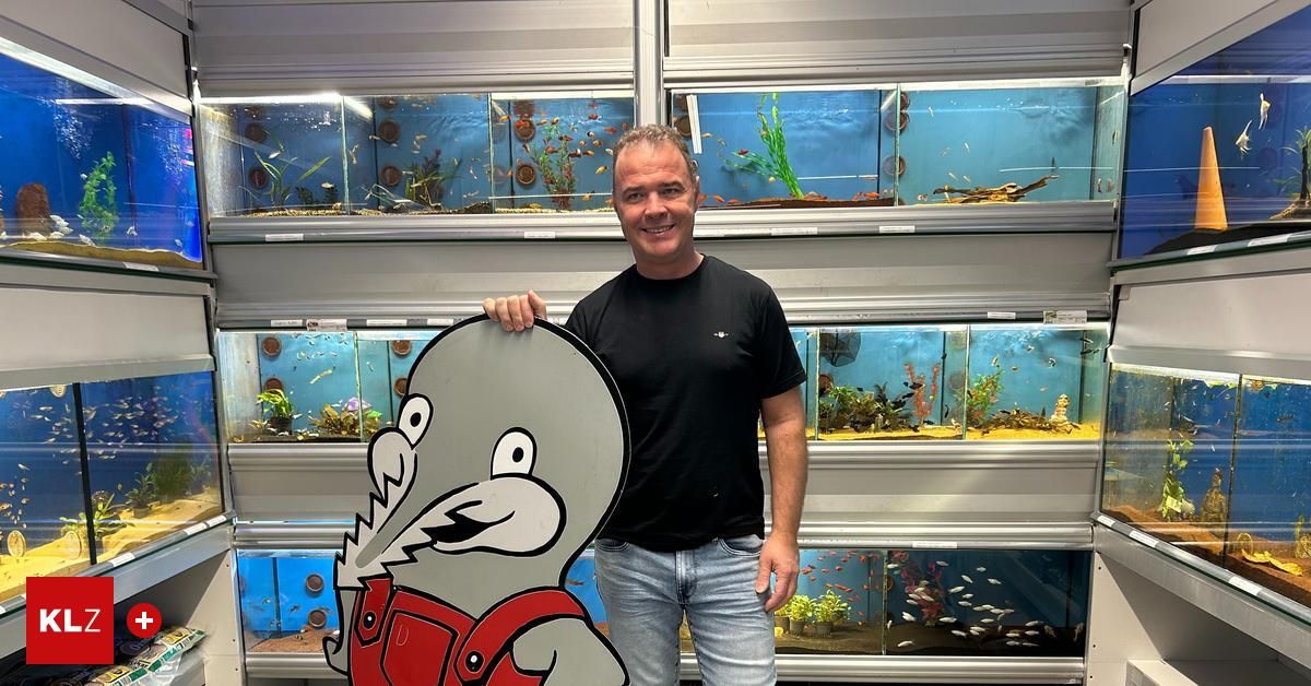 Ein Mann in einem schwarzen T-Shirt und Jeans lächelt und hält ein Schild mit einem gezeichneten Fisch in roten Overalls. Hinter ihm befinden sich mehrere Aquarien mit Fischen und Pflanzen.
