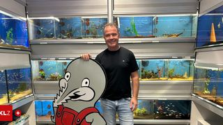 Ein Mann in einem schwarzen T-Shirt und Jeans lächelt und hält ein Schild mit einem gezeichneten Fisch in roten Overalls. Hinter ihm befinden sich mehrere Aquarien mit Fischen und Pflanzen.