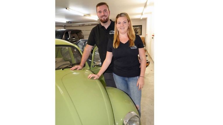 Ein Mann und eine Frau stehen in einer Garage neben einem grünen Oldtimer, beide tragen schwarze T-Shirts. Hinter ihnen sind mehrere Autos geparkt, und die Decke hat Leuchtstofflampen.