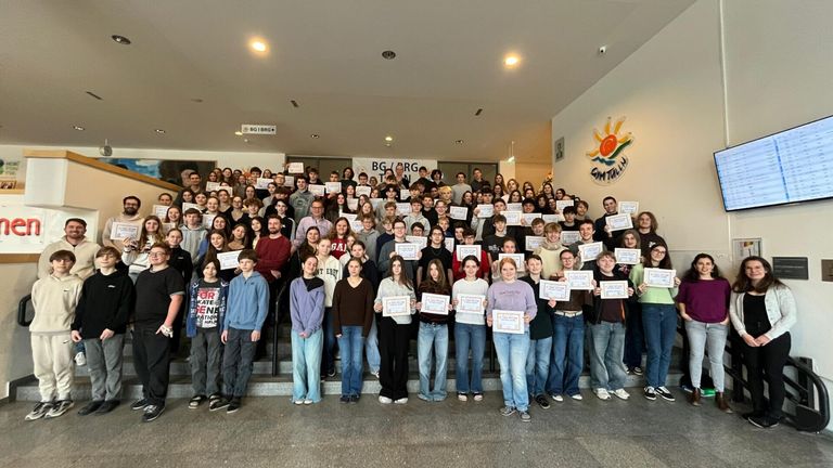 Eine große Gruppe von Menschen in Freizeitkleidung, die in Reihen stehen, Urkunden halten und lächelnd für ein Foto in einem Saal posieren.