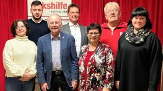 Fünf Personen, darunter ein Mann in einem marineblauen Anzug und eine Frau in einer Bluse mit Blumenmuster, stehen vor einem roten Vorhang. Dahinter steht ein Schild mit der Aufschrift 'kshilfe Poysdorf.'